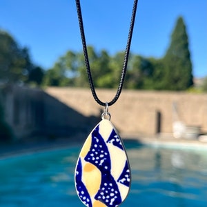 May include: A teardrop-shaped ceramic pendant necklace with a blue and yellow abstract pattern. The pendant is hanging from a black cord.