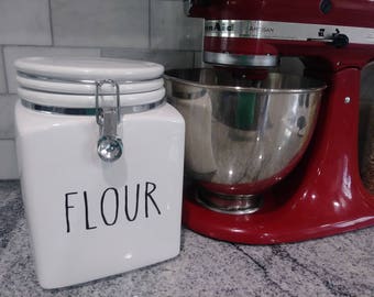 May include: White ceramic canister with a silver lid and the word "FLOUR" written in black. The canister is next to a red stand mixer with a silver bowl.