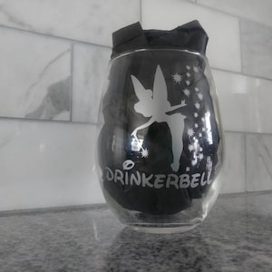 May include: A clear glass wine glass with a black and white design of Tinkerbell, a fairy with wings, and the text "DRINKERBELL".