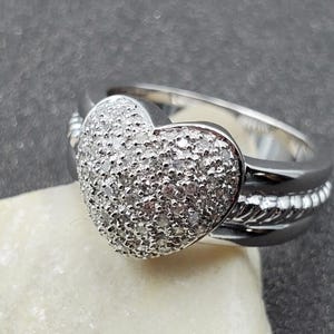 May include: A silver ring with a heart-shaped design. The heart is covered in small, sparkling stones. The ring has a braided band and a smooth, polished finish. A romantic piece of jewelry.