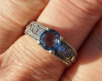 May include: A silver ring with a blue gemstone surrounded by smaller blue gemstones. The ring has an intricate design with a textured band.
