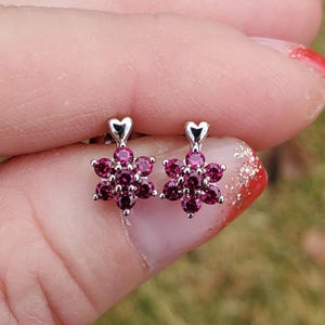 May include: A pair of silver earrings with a heart-shaped design and a cluster of pink gemstones in the shape of a flower.