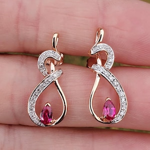 May include: A pair of rose gold earrings with a teardrop-shaped ruby gemstone and a pave setting of smaller clear stones.