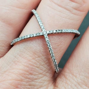 May include: A silver-toned ring with a crisscross design, encrusted with small, clear gemstones. The ring is worn on a finger, showcasing the intricate details of the jewelry.