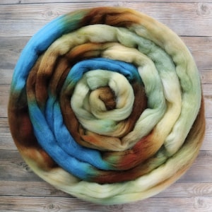Könnte beinhalten: Eine kreisförmige Spule aus weichem, ungesponnenem Wollvlies mit einer bunten Farbpalette aus Himmelblau, erdigem Braun, Creme und blassem Salbeigrün. Die Fasern sind in einem durchgehenden Spiralmuster angeordnet und zeigen eine Mischung aus Naturtönen.