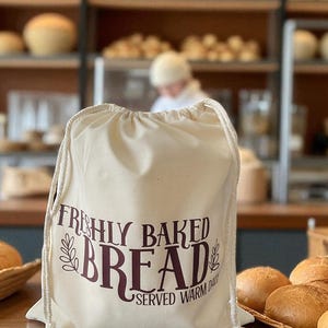 May include: A cream-coloured drawstring bag with the words "FRESHLY BAKED BREAD SERVED WARM DAILY" in burgundy lettering. The bag is on a wooden surface with baked goods in the background, suggesting a bakery setting.