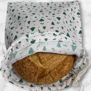 May include: A white bread bag with a drawstring closure, patterned with green Christmas trees and red accents. The bag contains a round loaf of bread with a golden-brown crust. The bag is made of fabric.