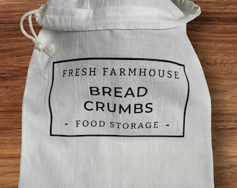 May include: A white fabric bag with a drawstring closure, labeled "FRESH FARMHOUSE BREAD CRUMBS - FOOD STORAGE" in black text. The bag is filled with bread crumbs and sits on a wooden surface.