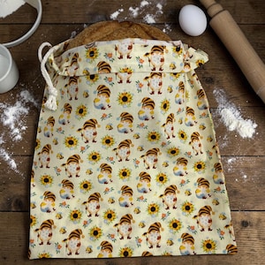 May include: A beige bread bag with a drawstring closure, filled with a loaf of bread. The bag is patterned with cartoon gnomes, sunflowers, and bees. The bag is made of fabric and has a white drawstring.