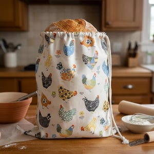 May include: A white fabric drawstring bread bag featuring a repeating pattern of colorful chickens in various styles and colors, filled with a round loaf of crusty bread. The bag has a thick white cord closure at the top.