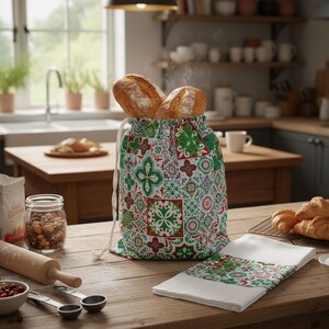 May include: A fabric drawstring bread bag featuring a colourful red, green, and white geometric tile pattern, filled with two crusty loaves of bread. A matching white kitchen towel with a patterned border is folded on the wooden surface nearby.