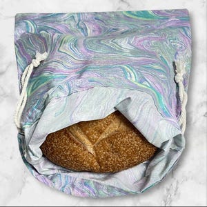 May include: A reusable bread bag with a marbled pattern in shades of purple, teal, and white. The bag is open, revealing a loaf of crusty, golden-brown bread. The bag has a drawstring closure made of white rope.