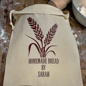 May include: A beige canvas bread bag with a drawstring closure. The bag features a brown graphic of wheat stalks and the text "HOMEMADE BREAD BY SARAH" in brown lettering. The bag is sitting on a wooden surface.