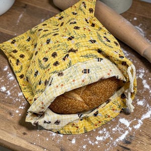 May include: A yellow fabric bread bag featuring a repeating pattern of bees, pinecones, and evergreen sprigs on a light yellow gingham background. The bag has a drawstring closure with white cord and contains a round, crusty loaf of bread.