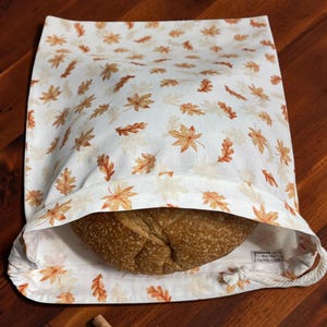 May include: A white fabric bread bag featuring a repeating pattern of orange, brown, and cream-coloured autumn leaves. The bag has a drawstring closure with white rope and contains a round loaf of crusty brown bread inside the open top.