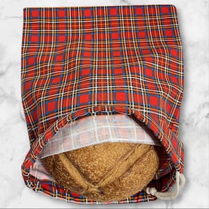 May include: A red plaid bread bag with a drawstring closure, holding a round loaf of bread. The bag's pattern includes red, blue, yellow, and white lines. The bread has a textured, golden-brown crust.