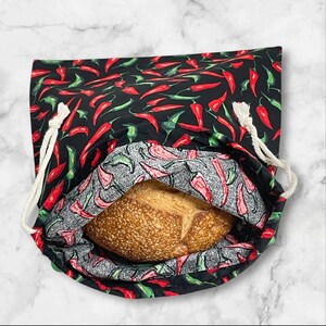 May include: A black bread bag with a red and green chilli pepper pattern. The bag is open, revealing a loaf of bread inside. The bag has a drawstring closure made of white rope.