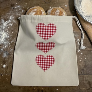 May include: A beige cotton drawstring bag featuring three vertically aligned red and white gingham hearts. The bag is positioned with two round loaves of bread tucked inside the top opening, with white flour scattered across the surface.