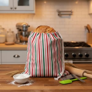 May include: A fabric bread bag featuring a vertical striped pattern in red, green, and white. The bag has a white drawstring closure at the top and contains a round loaf of crusty bread. The bag is made of a woven textile material.