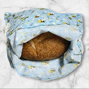 May include: A light blue bread bag with a drawstring closure, filled with a loaf of bread. The bag is patterned with yellow and black bees and white daisies. The bread has a golden-brown crust.