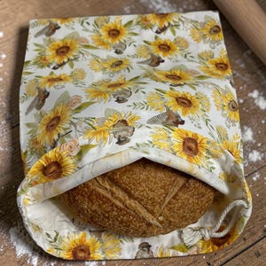May include: A bread bag with a sunflower pattern, holding a loaf of bread. The bag is white with yellow sunflowers and green leaves. A wooden rolling pin is in the background. The bread is brown and has a scored top.