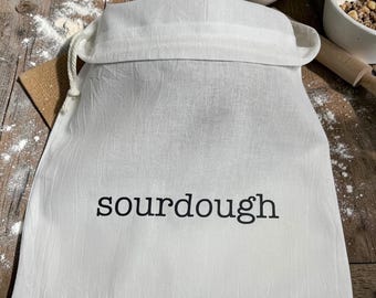May include: A white fabric drawstring bag featuring the word sourdough printed in black lowercase letters on the front. The bag is made of a textured, woven material and includes a thick white cord closure at the top for securing the contents.