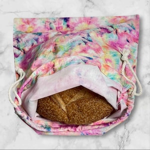 May include: A colourful tie-dye bread bag in pink, blue, yellow, and purple, with a drawstring closure. A loaf of brown bread is inside. The bag is made of fabric.