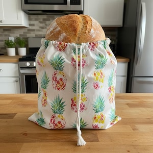 May include: A round loaf of bread sits inside a white drawstring bread bag. The bag is decorated with a repeating pattern of colorful pineapples in red, yellow, and green. The bag is tied with a white drawstring.