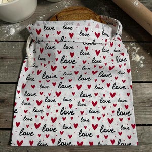 May include: A white fabric drawstring bag featuring a repeating pattern of small red hearts and the word love written in a black cursive script. The bag is partially filled with a round loaf of crusty brown bread, visible at the top opening.