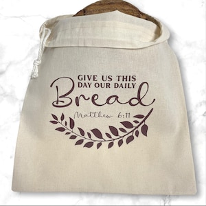 May include: A beige bread bag with a drawstring closure, holding a loaf of bread. The bag features the text "GIVE US THIS DAY OUR DAILY Bread" with "Matthew 6:11" below, and a decorative leaf design.