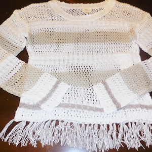 May include: White and beige striped sweater with long sleeves and fringe detail. The open-knit design features horizontal stripes of beige and white, with a fringe hemline. A casual, lightweight top.