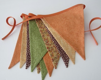 May include: A colorful fabric banner with triangular pennants in shades of orange, green, tan, and brown. The banner is trimmed with orange fabric and has orange ribbon ties on each end. The pennants feature various patterns, including polka dots and leaf designs.