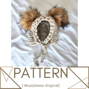 Può includere: Un berretto per neonato di colore bianco e beige lavorato a maglia con due pompon in pelliccia sintetica marrone. Il berretto ha un laccio sul retro. Il berretto è su una superficie bianca. [WoollyGems Original]