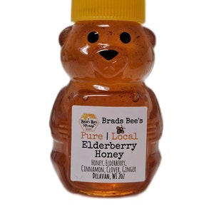 May include: A clear glass bear-shaped bottle filled with honey. The bottle has a yellow lid and a label that reads "Brads Bee's Honey Pure Local Elderberry Honey Honey, Elderberry, Cinnamon, Clover, Ginger Delavan, WI 202".