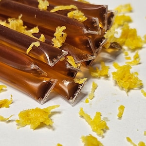 May include: A close-up of a pile of honey sticks, each individually wrapped in clear plastic. The sticks are filled with a golden honey liquid and are scattered on a white surface. There are small pieces of yellow citrus zest scattered around the honey sticks.