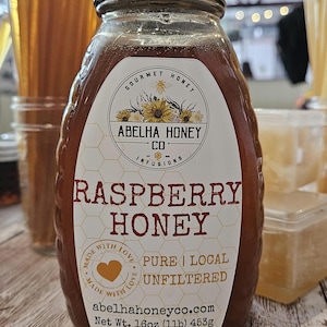 May include: A jar of raspberry honey with a silver lid and a white label. The label reads "Raspberry Honey" and "Pure, Local, Unfiltered." The jar is labeled "Abelha Honey Co." and contains 16oz (1lb) or 453g of honey. Honey sticks are visible in the background.
