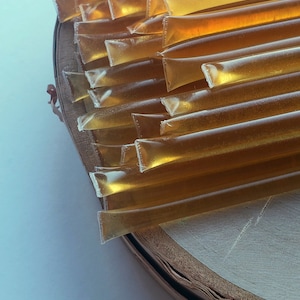 May include: A close-up of a stack of honey sticks on a wooden surface. The sticks are translucent and golden brown in color.