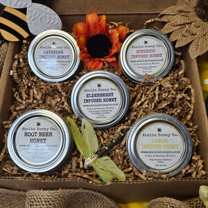 May include: A gift box filled with six small jars of honey infused with different flavors. The flavors are Lavender, Hibiscus, Elderberry, Root Beer, and Lemon. Each jar has a label with the Abelha Honey Co. logo and website address.