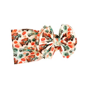 May include: A fabric headband featuring a large bow and a repeating pattern of teal vintage pickup trucks carrying orange pumpkins on a white background. The material has a textured, ribbed appearance throughout the entire accessory.