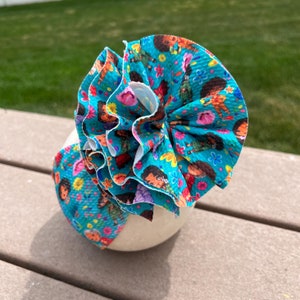 May include: A teal baby headband with a large, ruffled bow. The bow and headband are patterned with cartoon characters, flowers, and other colorful elements. The bow is layered with white fabric.