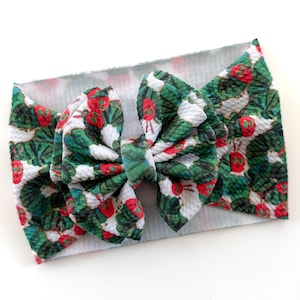 May include: A fabric headband with a large bow. The bow and headband feature a repeating pattern of green leaves and red berries on a white background. The bow is centered on the headband.