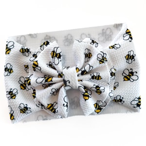 May include: White baby headband with a large bow featuring a repeating pattern of black and yellow bees. The headband is made of a soft, textured fabric, perfect for a baby girl.
