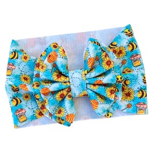 May include: A baby headband with a large bow featuring a blue background with sunflowers, honey pots, and bees. The bow is made of a soft, textured fabric. The headband is designed for infants and toddlers.