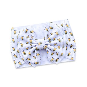 May include: White baby headband with a large bow featuring a repeating pattern of yellow and black bees. The headband is made of a soft, textured fabric, perfect for infants and toddlers. The bow is centered on the headband.