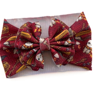 May include: A burgundy baby headband with a large bow featuring a repeating pattern of owls, books, and other magical elements. The headband is a soft, stretchy fabric, perfect for newborns and infants.