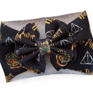 May include: Black headband with a large bow featuring a repeating pattern of gold text and white symbols. The design includes the words "Potter" and the Deathly Hallows symbol. The center of the bow has a crest design.