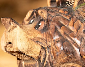 May include: A hand-carved wooden bear sculpture featuring a dark brown finish with visible chisel marks. The bear is depicted in profile, showing a textured snout, rounded ears, and painted eyes, with a focus on the detailed wood grain.