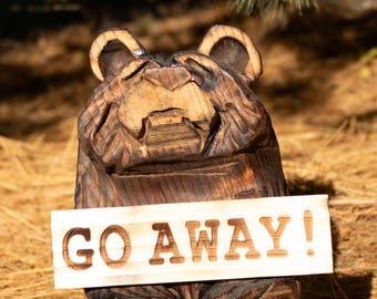 May include: A dark brown, hand-carved wooden bear sculpture with a textured finish. The bear is sitting upright and holding a light-colored rectangular wooden sign that features the text GO AWAY! carved in bold, dark capital letters.