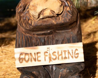 May include: A dark brown, hand-carved wooden bear sculpture with a light-colored wooden sign attached to its chest. The sign features the engraved text GONE FISHING with a small fish icon replacing the letter I in the word FISHING.