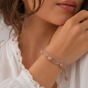 May include: A macrame bracelet with peach-colored beads. The bracelet features a woven design with small metallic accents. The beads are evenly spaced along the bracelet. The bracelet is on a wrist.